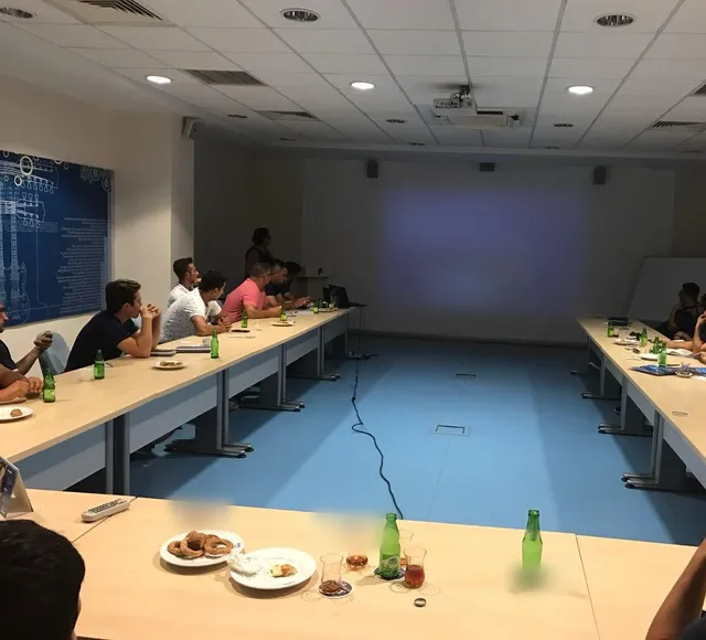 U düzenindeki toplantı odasında perdeye yansıtılan sunumu izleyen katılımcılar